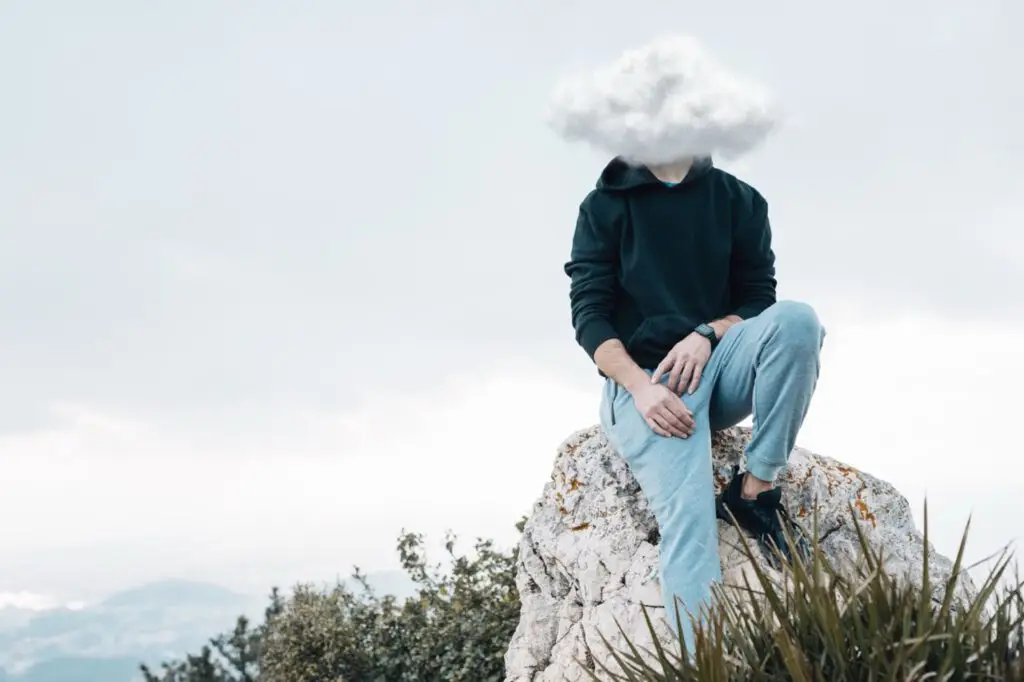 Person sitzt auf einem Felsen am Meer, eine Wolke verdeckt den Kopf als Sinnbild für Overthinking