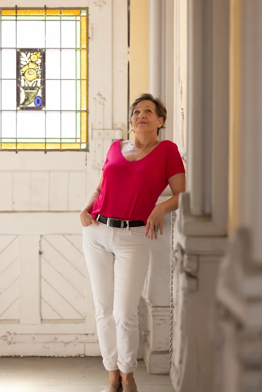 Andrea Däberitz lehnt lächelnd im roten Oberteil an einer Säule in einem hellen Saal