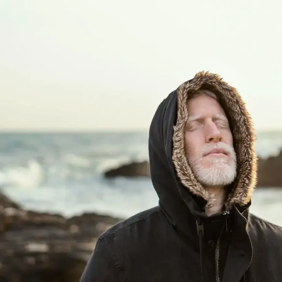 Mann mit Kapuze steht mit geschlossenen Augen am Meer